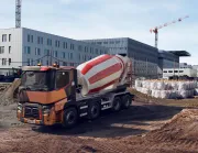 A Renault Trucks C lorry fitted with a concrete mixer is operating on a large urban construction site surrounded by cranes and modern buildings.